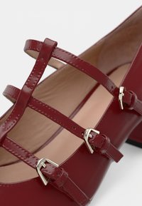 Red patent leather shoe with multiple strappy buckle accents and a rounded toe. Smooth texture and a slight heel.