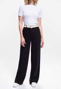 Donna che indossa una semplice t-shirt bianca a maniche corte infilata in pantaloni larghi neri con una cintura beige e sneaker bianche.