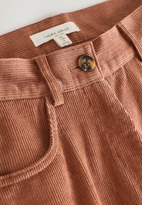 Rostes värvitud kordsest kangast püksid soonilise tekstuuriga, millel on üksik nööpkinnitus ning pistikdetailid taskutel ja vöökohal.