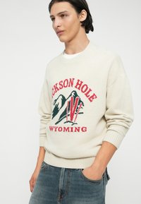Crème trui met geribbelde manchetten en onderkant, met een kleurrijk gebreid ontwerp van bergen en de tekst "JACKSON HOLE WYOMING" in rood en groen.