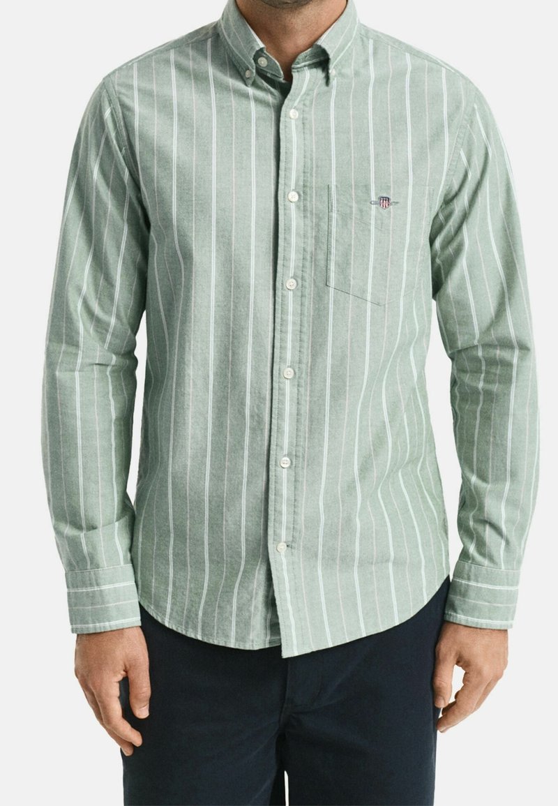 Homme portant une chemise à manches longues vert clair à rayures verticales blanches et un petit logo sur la poche.