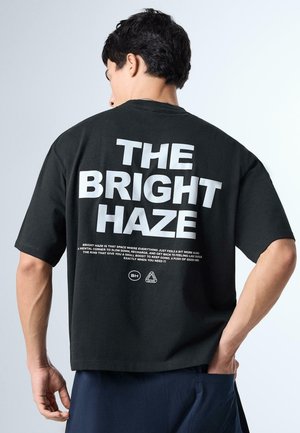 Muž s krátkymi čiernymi vlasmi má na sebe čierne tričko s veľkým bielym nápisom "THE BRIGHT HAZE" a menším textom pod ním na chrbte.
