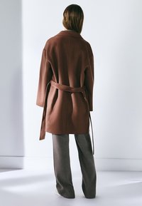 Cappotto in lana marrone con vita cintata, maniche lunghe e vestibilità rilassata. Abbinato a pantaloni in maglia grigia. Lo sfondo è un interno minimalista.