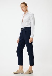 Vit skjorta med knappar, marinblå byxor med bälte i midjan och beige skor utan snörning. Slät tyg och skräddarsydd passform. Enkel design, neutrala färger.