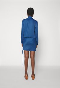 Satin blaues Kleid mit einer kragenlosen Jacke und einem Zugband-Detail an der Seite. Mit langen Ärmeln und texturalen Akzenten. Kräftige mehrfarbige Absätze.