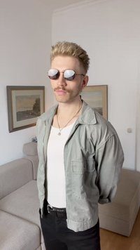 Lys grå zip-up jakke over en hvid skjorte, sorte bukser og runde solbriller med spejlelinser, stående i en simpel indendørs setting.