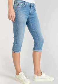 Pantalons capri en denim bleu clair avec ourlet retroussé, dotés de cinq poches et d'un design ajusté, associés à des baskets blanches.