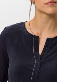 Marineblaues Oberteil mit Rundhalsausschnitt, langen Ärmeln und einer silbernen Akzentnaht entlang der Knopfleiste, gefertigt aus glattem Material.