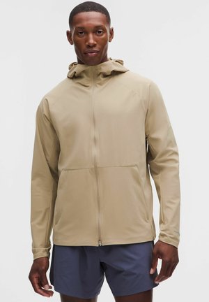 Hombre con chaqueta con capucha beige con cremallera y pantalones cortos azul oscuro de pie frente a un fondo claro y liso.