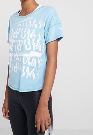Persoon die een lichtblauw Puma T-shirt met witte tekst draagt en zwarte leggings met een wit Puma-logo aan de zijkant, licht naar links gewend.