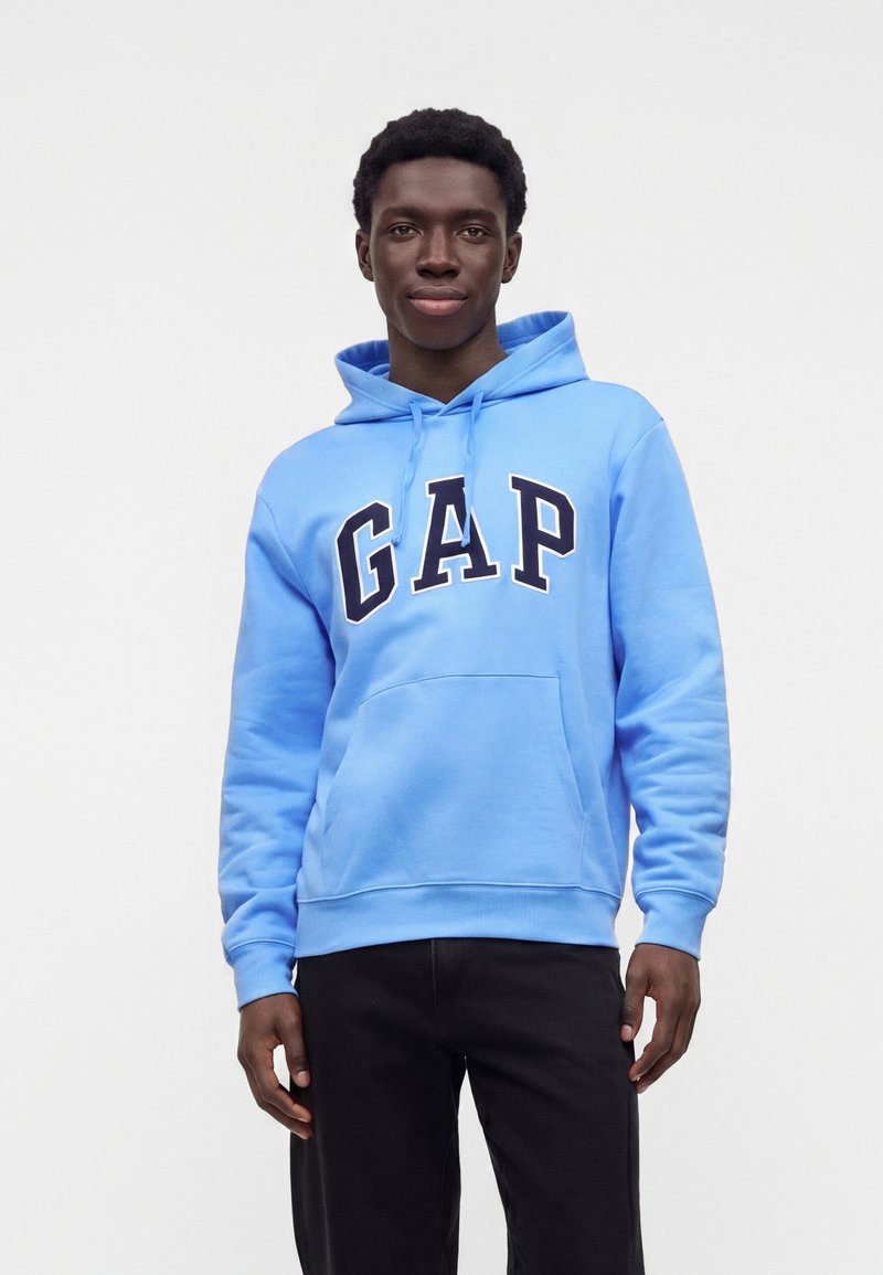 Jeune homme portant un sweat à capuche GAP bleu clair et un pantalon noir, debout devant un fond clair uni.