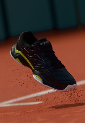 Zapatilla deportiva negra con acentos amarillos, superficie texturizada y logotipo prominente. Situada en el aire, rodeada de partículas de polvo.