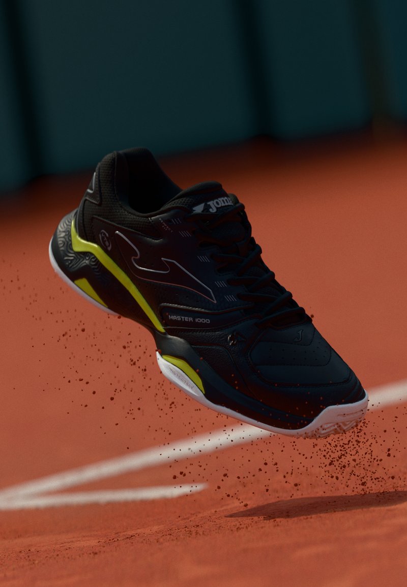 Shoe atlético preto com acentos amarelos, superfície texturizada e logotipo em destaque. Posicionado no ar, com partículas de poeira ao redor.