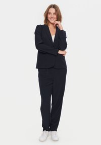 Jūras spēku krāsas vīriešu uzvalks ar vienu pogu un taisnām biksēm, apvienots ar baltiem sporta apaviem. Gluda auduma, minimāla dizaina, strukturēta piegriezuma.