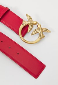 Ceinture en cuir rouge avec une boucle ronde en or en forme de deux oiseaux, avec des gravures détaillées. La ceinture comprend plusieurs trous pour l'ajustement.