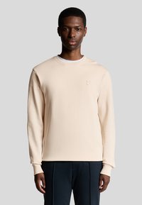 Licht beige crewneck sweatshirt van zachte stof met lange mouwen. Voorzien van een geborduurd logo op de borst, gecombineerd met donkere broeken.
