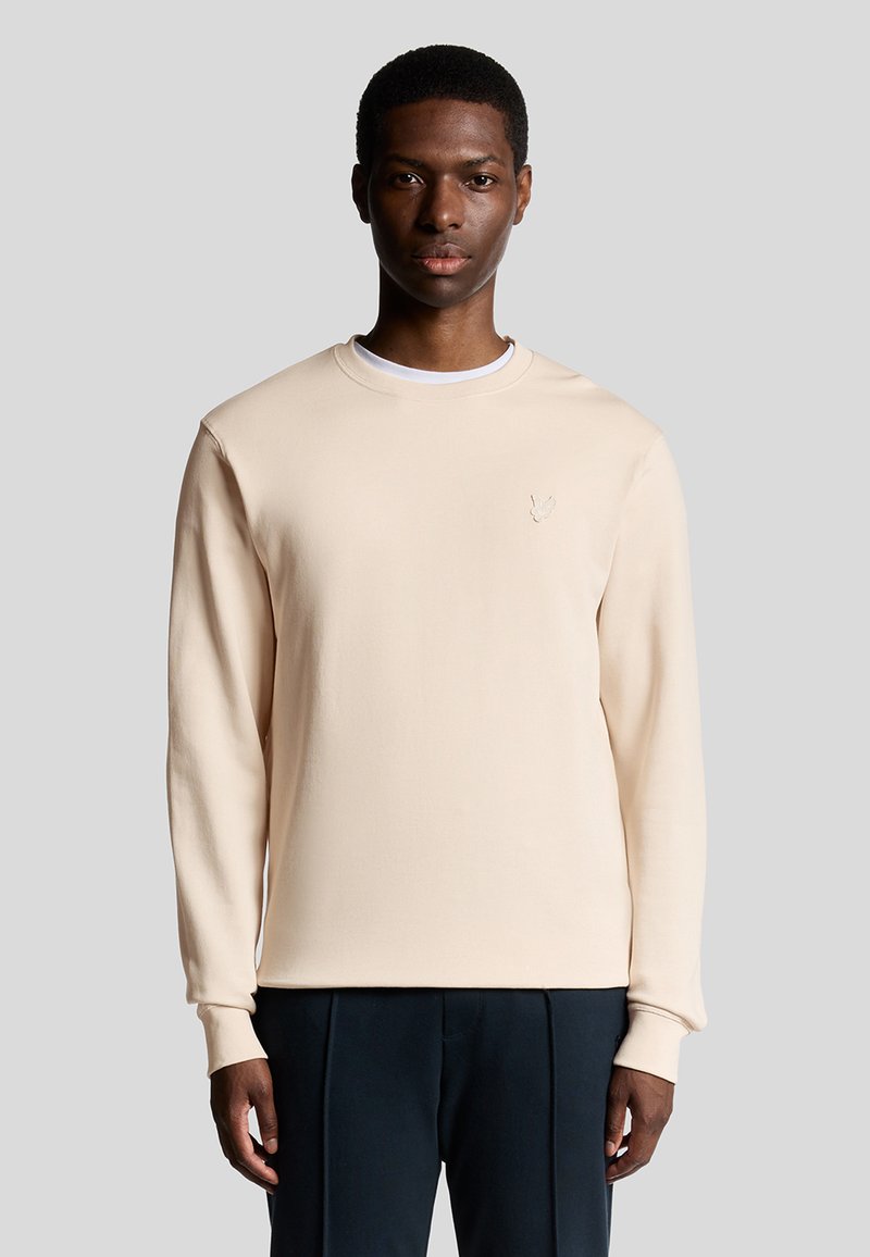 Licht beige crewneck sweatshirt van zachte stof met lange mouwen. Voorzien van een geborduurd logo op de borst, gecombineerd met donkere broeken.