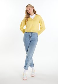 Cardigan giallo con scollo a V e chiusura a bottoni, abbinato a jeans a vita alta di colore blu chiaro. Presenta maniche arrotolate e sneaker multicolori.