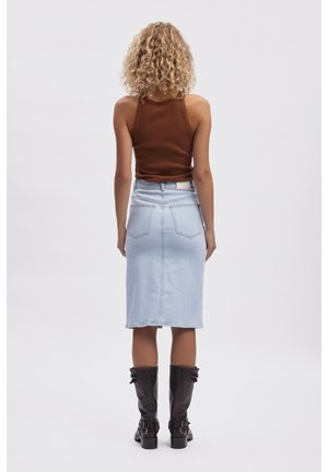 Kvinde med krøllet blondt hår iført en brun ærmeløs top, lyseblåt denimskaørt og sorte midt-lægstøvler, står med ryggen mod.