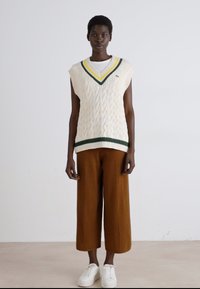 Pull en mailles blanches sans manches avec un col en V vert et jaune, associé à un pantalon large marron et des baskets blanches. Texture lisse.