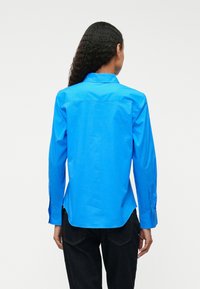 Sinine pika varrukaga särk, mis on valmistatud siledast kangast, koos krae ja nööpidega mansettidega, kaardus serv ja minimaalsete disainidetailidega.