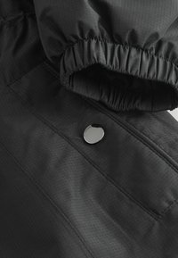 Manche de veste noire avec poignet élastique et bouton-pression métallique sur tissu avec détail de couture visible.