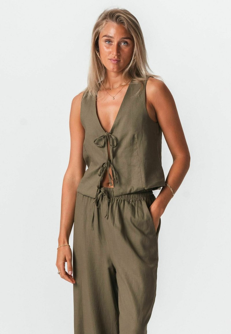 Femme aux cheveux blonds portant un haut sans manches noué devant de couleur vert olive et un pantalon ample assorti, debout avec une main dans la poche.