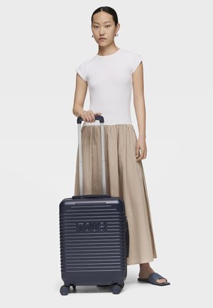 Giovane donna con top bianco e gonna beige in piedi accanto a una valigia blu navy con ruote e manico esteso su sfondo bianco.