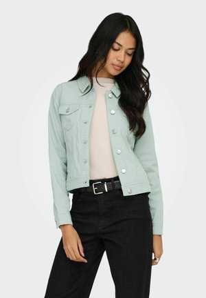 Mlada ženska, oblečena v svetlo zeleno jakno z gumbi, bež majico, črni pas in črne kavbojke, ki pozira z nagnjeno glavo in zaprtimi očmi.