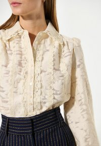 Blusa color crema con delicato pizzo, tessuto strutturato, bottoni al centro e maniche a sbuffo, abbinata a pantaloni a vita alta a righe blu navy.