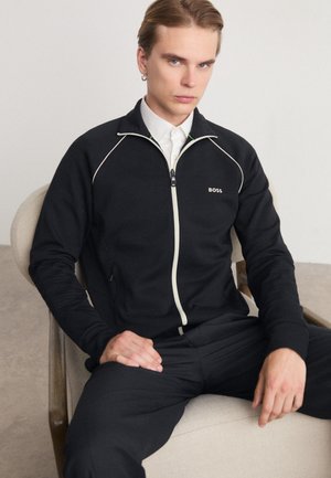 Veste zippée noire avec des finitions blanches et un petit logo sur la poitrine, fabriquée en tissu doux, associée à un pantalon foncé.