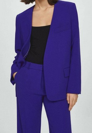Kvinde iført en kongeblå blazer og matchende bukser med en sort ærmeløs top, stående med den ene hånd i lommen mod en ensfarvet baggrund.