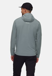 Lys blå hættetrøje med en tekstureret overflade, justerbar hætte og "MAMMUT" logo på ryggen. Designet til udendørsaktiviteter.