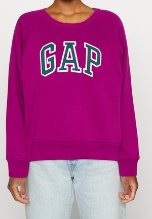 Personne portant un sweat-shirt violet vif avec le logo "GAP" en lettres blanches et vert foncé, combiné avec un jean en denim bleu clair.