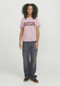 T-shirt di cotone rosa chiaro con "BARCELONA" stampato in rosso scuro, abbinata a jeans di denim blu larghi e zoccoli marroni, in piedi contro uno sfondo neutro.