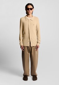 Beige texturerad kofta med krage och knappar, parat med lösa taupebyxor och bruna mockaskor. Mörka solglasögon fulländar looken.