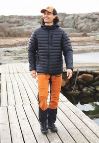 Svart puffjacka med en dragkedjeficka, orange och svarta byxor med dragkedjor, och svarta skor. Modellen står på en träbrygga vid vatten.