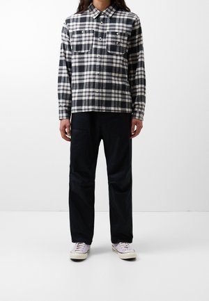 Persoon draagt een zwart-wit geruit overhemd met knopen, een zwarte broek en lichtpaarse sneakers, staand tegen een witte achtergrond.