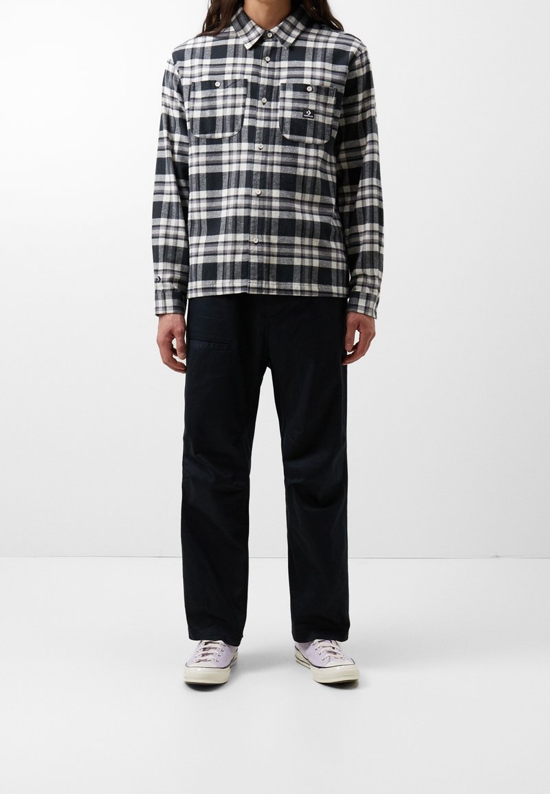 Personne portant une chemise à carreaux noire et blanche boutonnée, un pantalon noir et des baskets violet clair debout sur fond blanc.
