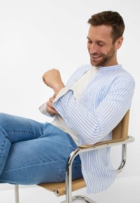 Camicia leggera a righe blu e bianche, con colletto alla mandarino, abbottonatura frontale e maniche lunghe arrotolate, abbinata a jeans blu.