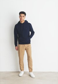 Marineblauwe sweatshirt met een capuchon en een voorzak, gecombineerd met beige broek. De outfit wordt compleet gemaakt met witte sneakers. Eenvoudig design.