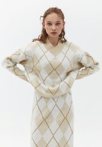 Beige- och vit rutmönstrad tröja med v-ringning och ribbade muddarna, tillverkad av ett mjukt, texturerat tyg. Diskreta metalliska detaljer överallt.