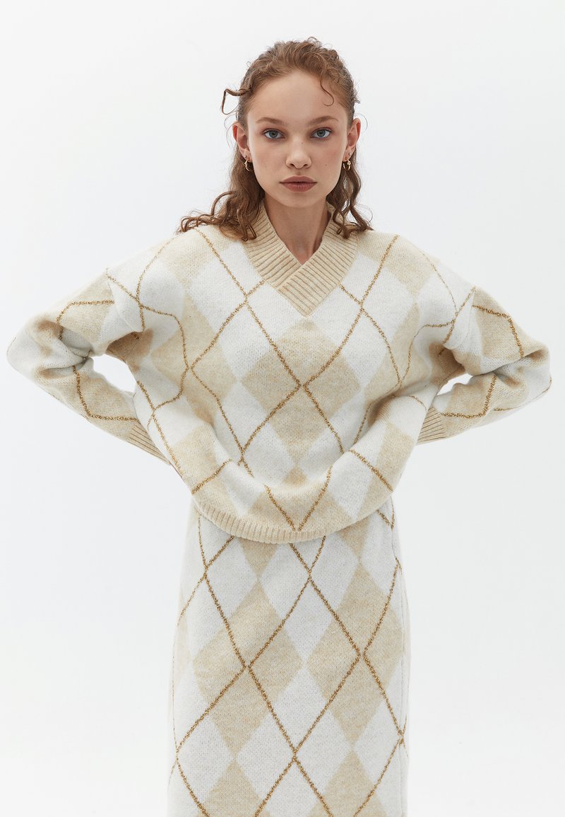 Beige- och vit rutmönstrad tröja med v-ringning och ribbade muddarna, tillverkad av ett mjukt, texturerat tyg. Diskreta metalliska detaljer överallt.