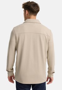 Camisa de manga larga beige con cuello, tejido texturizado y detalle en la parte trasera. Las aberturas laterales en el dobladillo mejoran la movilidad.