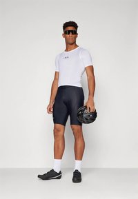 Bílé cyklistické triko s krátkým rukávem z síťoviny, černé cyklistické kraťasy, černé cyklistické boty a helma držená v jedné ruce, stojící před světlým pozadím.