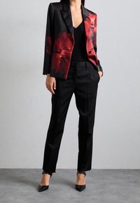 Schwarzer Blumenblazer mit roten Akzenten, kombiniert mit schwarzen taillierten Hosen und spitzen schwarzen Absätzen, vor einem neutralen Hintergrund.