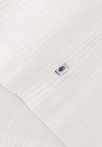 Witte textiele beddengoed met een subtiel geometrisch patroon. Bevat een klein stoffen label met een logo in een blauwe kleur.