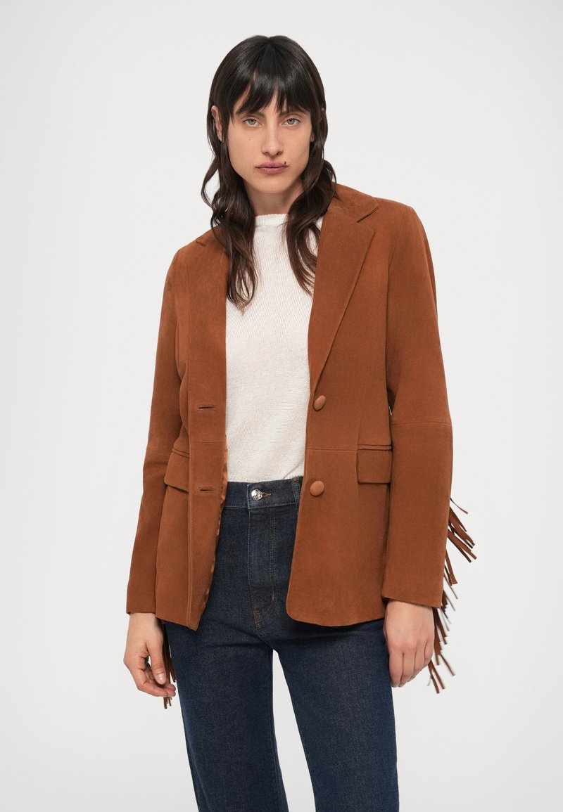 Brauner Wildlederblazer mit zwei vorderen Knöpfen, schmalen Revers und Seitentaschen. Verziert mit Fransen an den Ärmeln. Wird über einem leichten Pullover getragen.