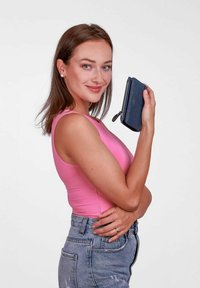 Námořnická modrá strukturovaná peněženka se zipem držena v ruce, s decentním logem. Model má na sobě růžový top a džíny.