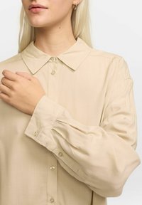 Beige Bluse mit Kragen, Knopfleiste und langen Ärmeln. Weicher Stoff mit leichtem Glanz und gerafftem Ärmeldetail.